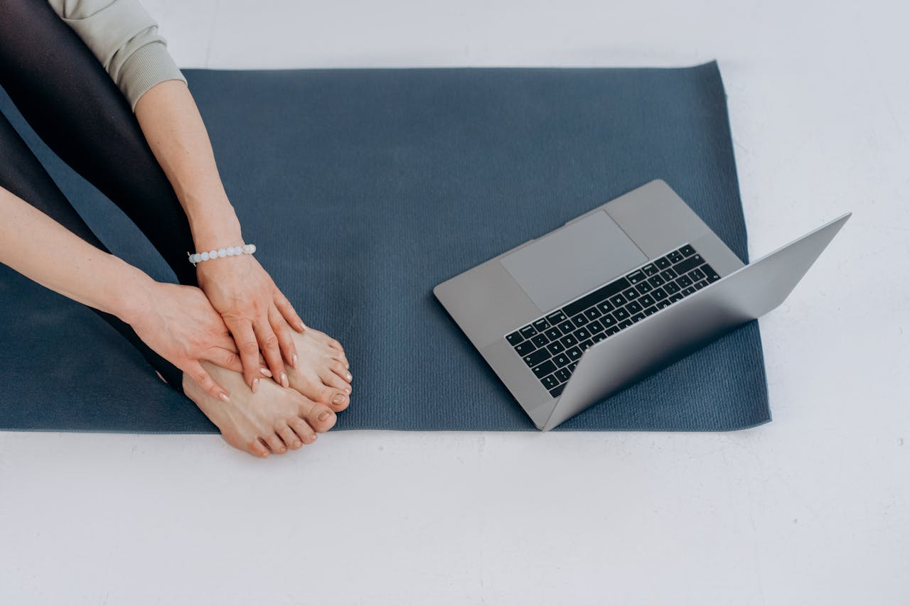 Individual on yoga mat practicing stretches alongside a laptop for online fitness classes.